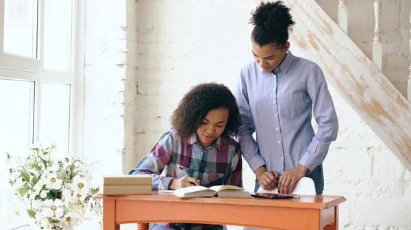 Junges Mädchen mit lockigem Haar und gemischter Rasse sitzt am Tisch und konzentriert sich auf den Unterricht und ihre ältere Schwester hilft ihr beim Lernen — Stockfoto