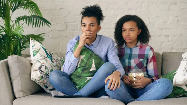 Zwei gemischte Rasse lockige Freundinnen sitzen auf der Couch und sehen Thriller-Film und essen Popcorn zu Hause — Stockfoto