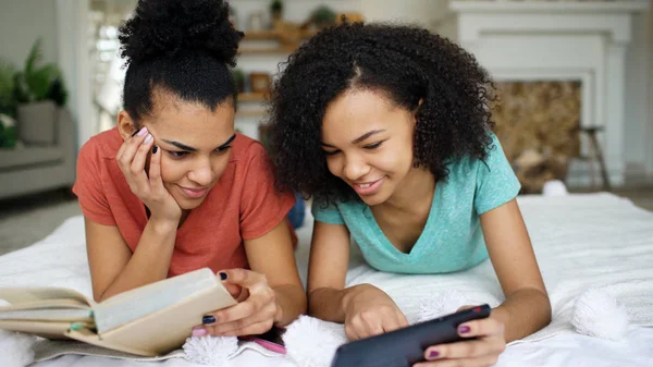 Two cheerful mixed race funny girlfriends reading book and using tablet computer talks and have fun lying in bed at home — Stock Photo, Image