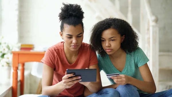 Two cheerful mixed race curly girlfriends shopping online with tablet computer and credit card at home — Stock Photo, Image