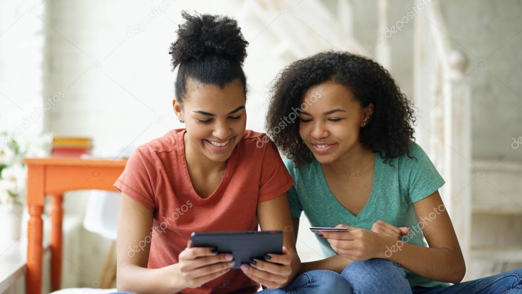 Two cheerful mixed race curly girlfriends shopping online with tablet computer and credit card at home