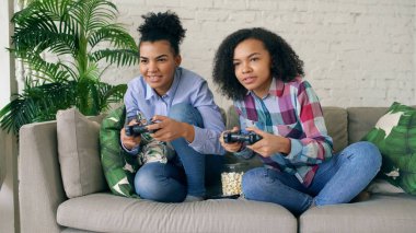 İki karma yarış kıvırcık kız kanepede oturan gamepad ile konsol bilgisayar oyunları oynamak ve evde eğlenmek
