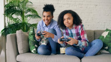 İki karma yarış kıvırcık kız kanepede oturan gamepad ile konsol bilgisayar oyunları oynamak ve evde eğlenmek