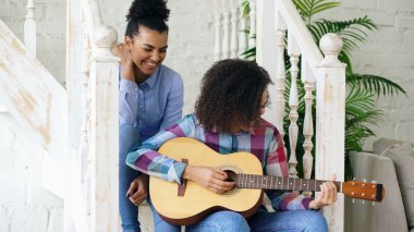 Merdiven ve akustik gitar play uygulama üzerinde oturan iki Brezilyalı kıvırcık kız sistres. Eğlenceli ve evde şarkı arkadaşlarım var.