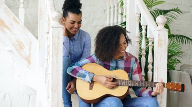 Merdiven ve akustik gitar play uygulama üzerinde oturan iki Brezilyalı kıvırcık kız sistres. Eğlenceli ve evde şarkı arkadaşlarım var.