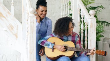 Merdiven ve akustik gitar play uygulama üzerinde oturan iki Brezilyalı kıvırcık kız sistres. Eğlenceli ve evde şarkı arkadaşlarım var.