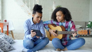 Akustik gitar evde oynamak için genç kız kardeşi öğretim yatağa oturmuş tablet bilgisayar ile karışık yarış genç kadın