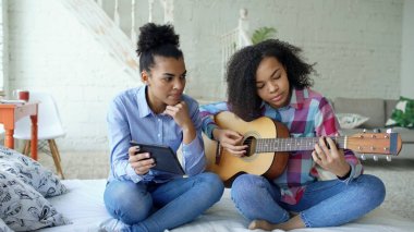 Akustik gitar evde oynamak için genç kız kardeşi öğretim yatağa oturmuş tablet bilgisayar ile karışık yarış genç kadın
