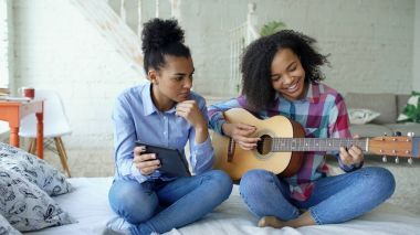 Akustik gitar evde oynamak için genç kız kardeşi öğretim yatağa oturmuş tablet bilgisayar ile karışık yarış genç kadın