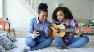 Akustik gitar evde oynamak için genç kız kardeşi öğretim yatağa oturmuş tablet bilgisayar ile karışık yarış genç kadın