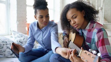 Akustik gitar evde oynamak için genç kız kardeşi öğretim yatağa oturmuş tablet bilgisayar ile karışık yarış genç kadın