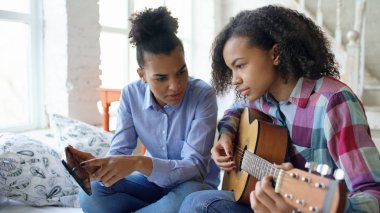 Akustik gitar evde oynamak için genç kız kardeşi öğretim yatağa oturmuş tablet bilgisayar ile karışık yarış genç kadın
