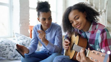 Akustik gitar evde oynamak için genç kız kardeşi öğretim yatağa oturmuş tablet bilgisayar ile karışık yarış genç kadın
