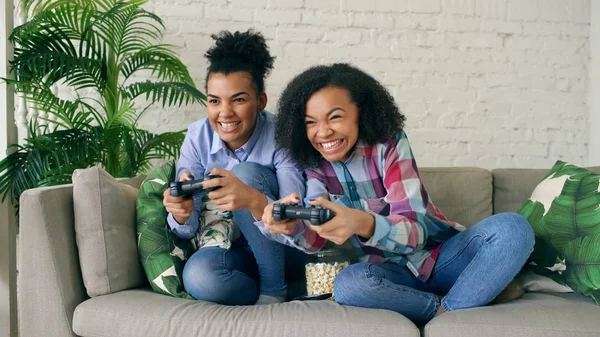 İki karma yarış kıvırcık kız kanepede oturan gamepad ile konsol bilgisayar oyunları oynamak ve evde eğlenmek
