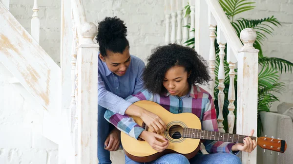 Mujer joven de raza mixta sentada en escaleras enseñando a su hermana adolescente a tocar la guitarra acústica en casa — Foto de Stock