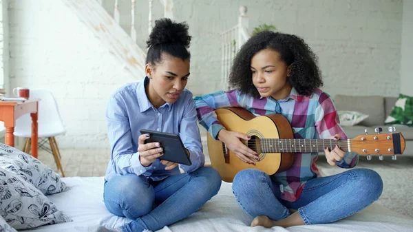 Змішана раса молода жінка з планшетним комп'ютером сидить на ліжку навчаючи її сестру-підлітка грати на акустичній гітарі вдома — стокове фото