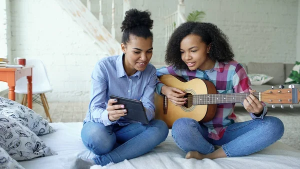 Змішана раса молода жінка з планшетним комп'ютером сидить на ліжку навчаючи її сестру-підлітка грати на акустичній гітарі вдома — стокове фото