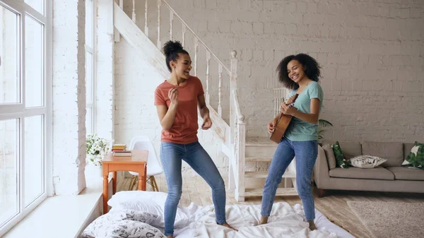 Mieszane rasy młode dziewczyny zabawne taniec, śpiew i gra na gitarze akustycznej na łóżku. Siostry, zabawy rekreacyjne w sypialni w domu — Zdjęcie stockowe