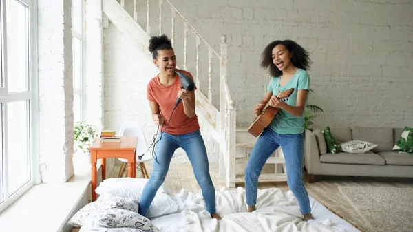 Raza mixta de chicas divertidas jóvenes bailan cantando con secador de pelo y tocando la guitarra acústica en una cama. Hermanas divertirse ocio en el dormitorio en casa — Foto de Stock