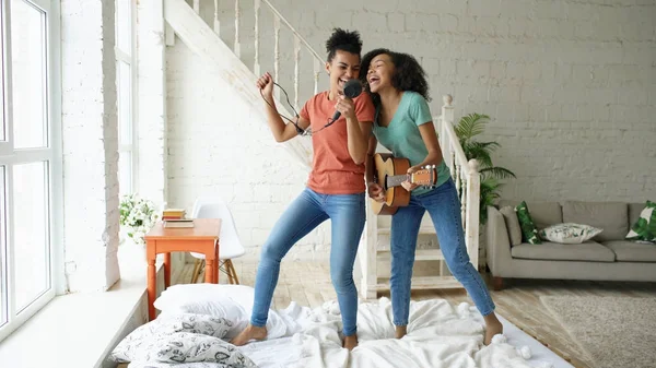 Młode dziewczyny śmieszne mieszane rasy taniec śpiewa z suszarką do włosów i gra gitara akustyczna na łóżku. Siostry, zabawy rekreacyjne w sypialni w domu — Zdjęcie stockowe
