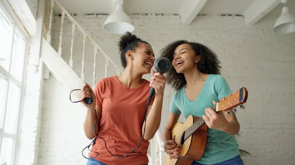 Raza mixta de chicas divertidas jóvenes bailan cantando con secador de pelo y tocando la guitarra acústica en una cama. Hermanas divertirse ocio en el dormitorio en casa — Foto de Stock