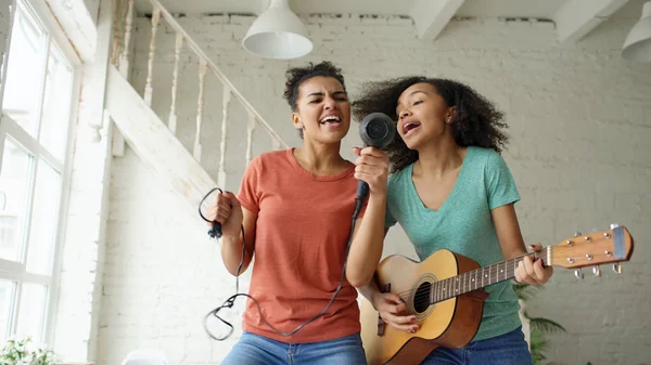 Смешанные расы юные смешные девушки танцуют с феном и играют на акустической гитаре на кровати. Систеры весело проводят время в спальне дома — стоковое фото