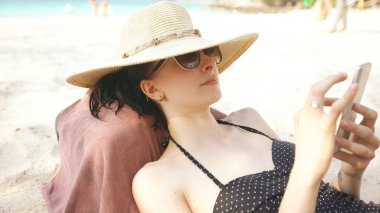 Şapka ve güneş gözlüğü smartphone ve yalanlar kumsalda kullanarak genç kadın