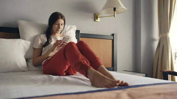 Όμορφη γυναίκα ξαπλωμένη στο κρεβάτι σε δωμάτιο ξενοδοχείου και χρησιμοποιώντας το smartphone και χαμογελαστός — Φωτογραφία Αρχείου