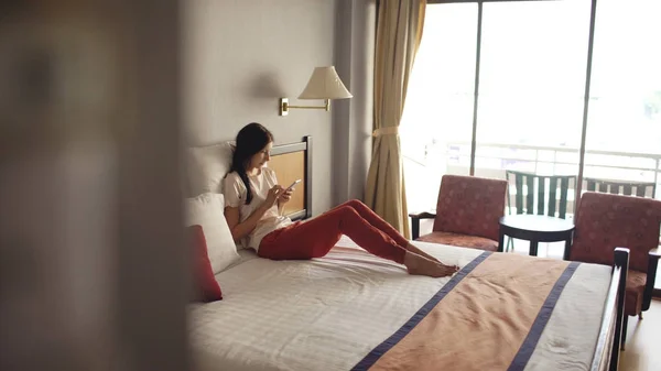 Mooie vrouw liggend in bed in een hotelkamer en het gebruik van smartphone en glimlachen — Stockfoto