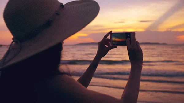 Силуэт молодой туристки в шляпе, фотографирующей с сотовым телефоном во время захода солнца на берегу океана — стоковое фото