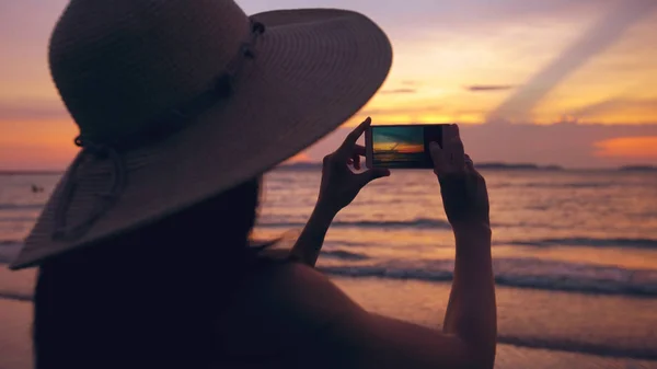 Силуэт молодой туристки в шляпе, фотографирующей с сотовым телефоном во время захода солнца на берегу океана — стоковое фото