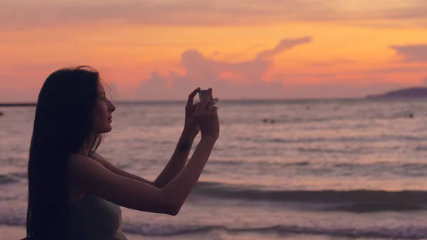 Młody turysta kobieta fotografie ocean widok ze smartfona podczas zachodu słońca na plaży — Zdjęcie stockowe