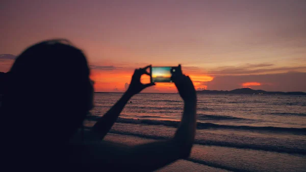 Крупный план молодой туристки фотографирует вид на океан со смартфона на закате на пляже — стоковое фото