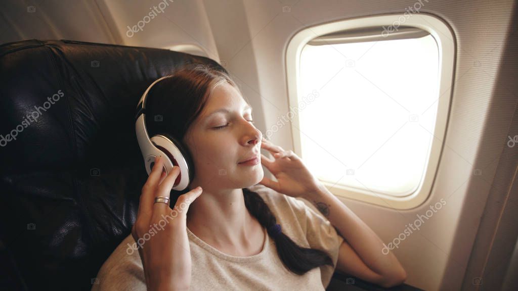 Young woman in wireless headphones listening to music and smiling during fly in airplane