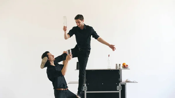 Hombres camareros profesionales haciendo malabarismos con botellas y cócteles temblorosos en la mesa de bar móvil en el estudio de fondo blanco en interiores — Foto de Stock