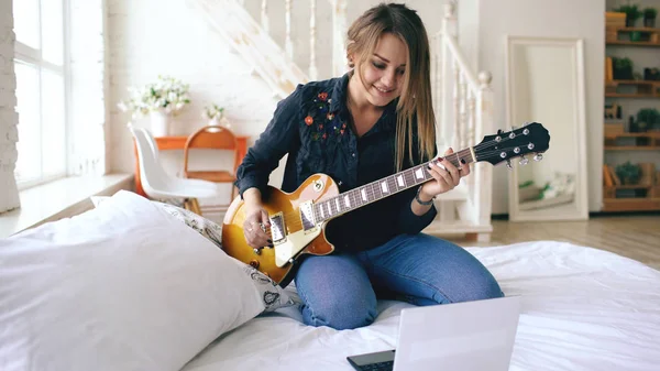 Atractiva niña aprendiendo a tocar la guitarra eléctrica con portátil sentarse en la cama en el dormitorio en casa — Foto de Stock