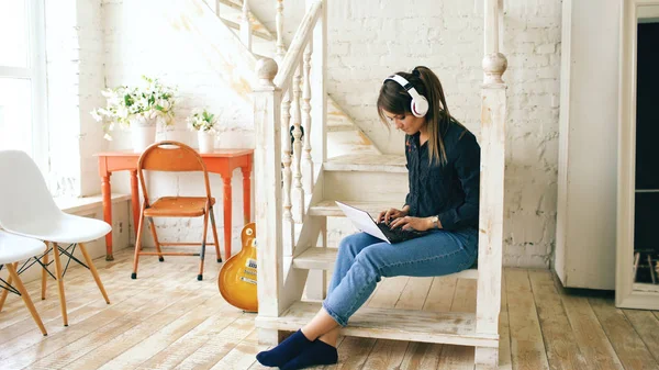 Hermosa mujer con auriculares y portátil posando y sonriendo mientras se sienta en las escaleras en casa —  Fotos de Stock