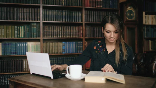 Estudante sorrindo menina trabalhando no laptop e ler livro na biblioteca da universidade dentro de casa — Fotografia de Stock