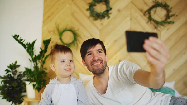 スマート フォンのカメラで selfie 写真を取ってベッドの上の息子と若い笑顔の父 — ストック写真
