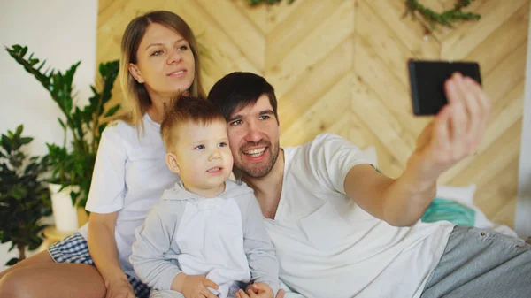 Pais sorridentes com bebê tirando foto da família selfie na cama em casa — Fotografia de Stock