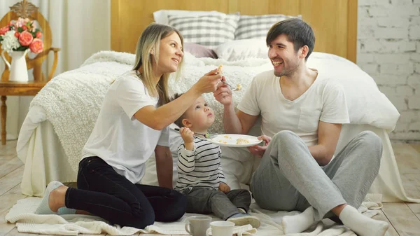Pequeño niño adorable celebrando su cumpleaños con padre y madre comer pastel en el dormitorio — Foto de Stock