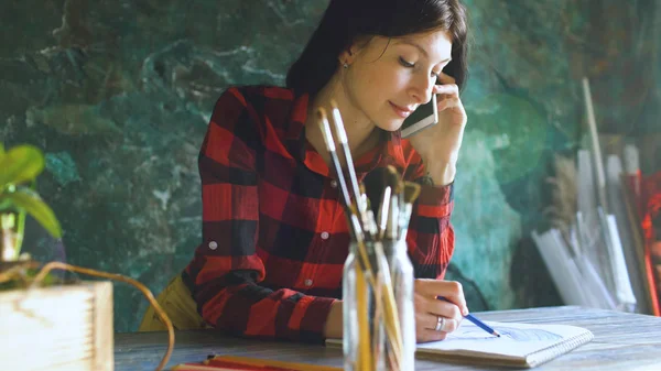 Молода жінка художник малює клаптик на паперовому ноутбуці з олівцем і розмовляє телефоном у приміщенні — стокове фото