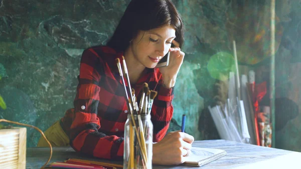 Jovem artista pintando scetch no caderno de papel com lápis e telefone falando dentro de casa — Fotografia de Stock
