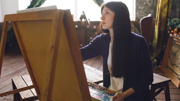 Молода жінка-художниця малює малювання акварельними фарбами та пензлем на мольберті — стокове фото