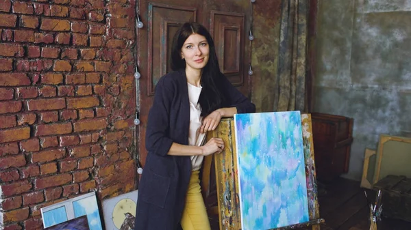 Giovane artista donna con vernici ad acquerello stand vicino immagine finita e sorriso in macchina fotografica — Foto Stock