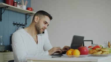 Telefonu konuşurken ve sabah kahvaltıdan sonra mutfakta otururken dizüstü bilgisayar kullanan yakışıklı delikanlı