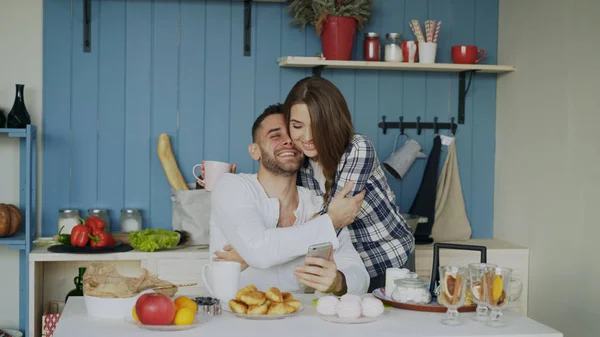 Atractiva pareja se reúnen en la cocina a la hora del desayuno y el uso de teléfonos inteligentes para navegar en las redes sociales en casa — Foto de Stock