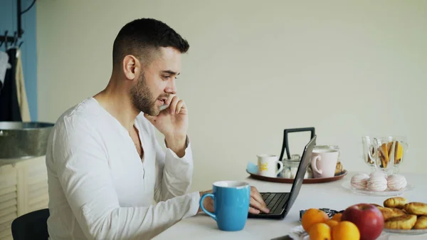 Hombre joven guapo hablando de teléfono y el uso de ordenador portátil sentado en la cocina después del desayuno por la mañana — Foto de Stock