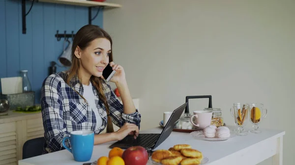 Dizüstü bilgisayar kullanan ve sabahları kahvaltı sırasında telefonu konuşurken sosyal medya tarama genç gülümseyen kadın — Stok fotoğraf