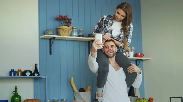 Boldog fiatal pár szórakozik otthon a konyhában. Lány ül a barátok nyak, miközben ő selfie portré véve a smartphone kamera — Stock Fotó